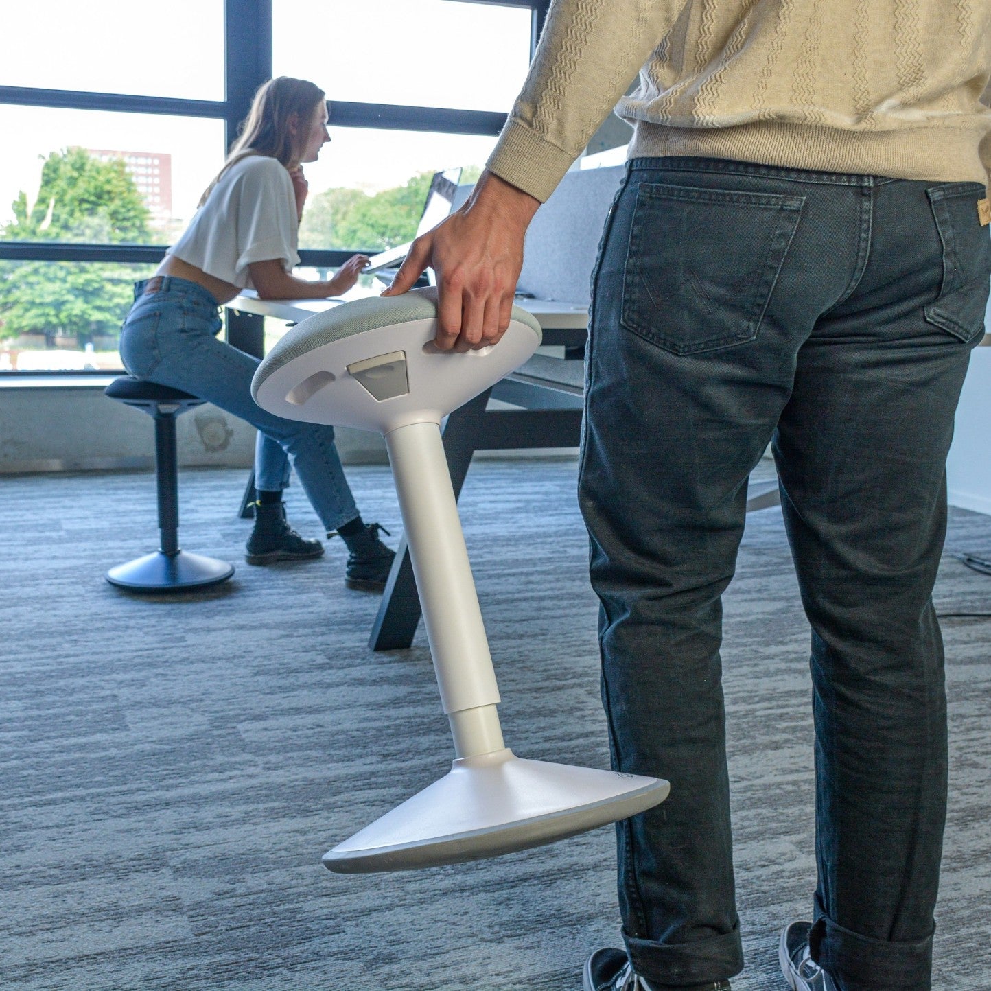 Een man in een kantoor met een laptop, terwijl hij naast een duurzame wiebelkruk staat die ergonomisch is ontworpen.